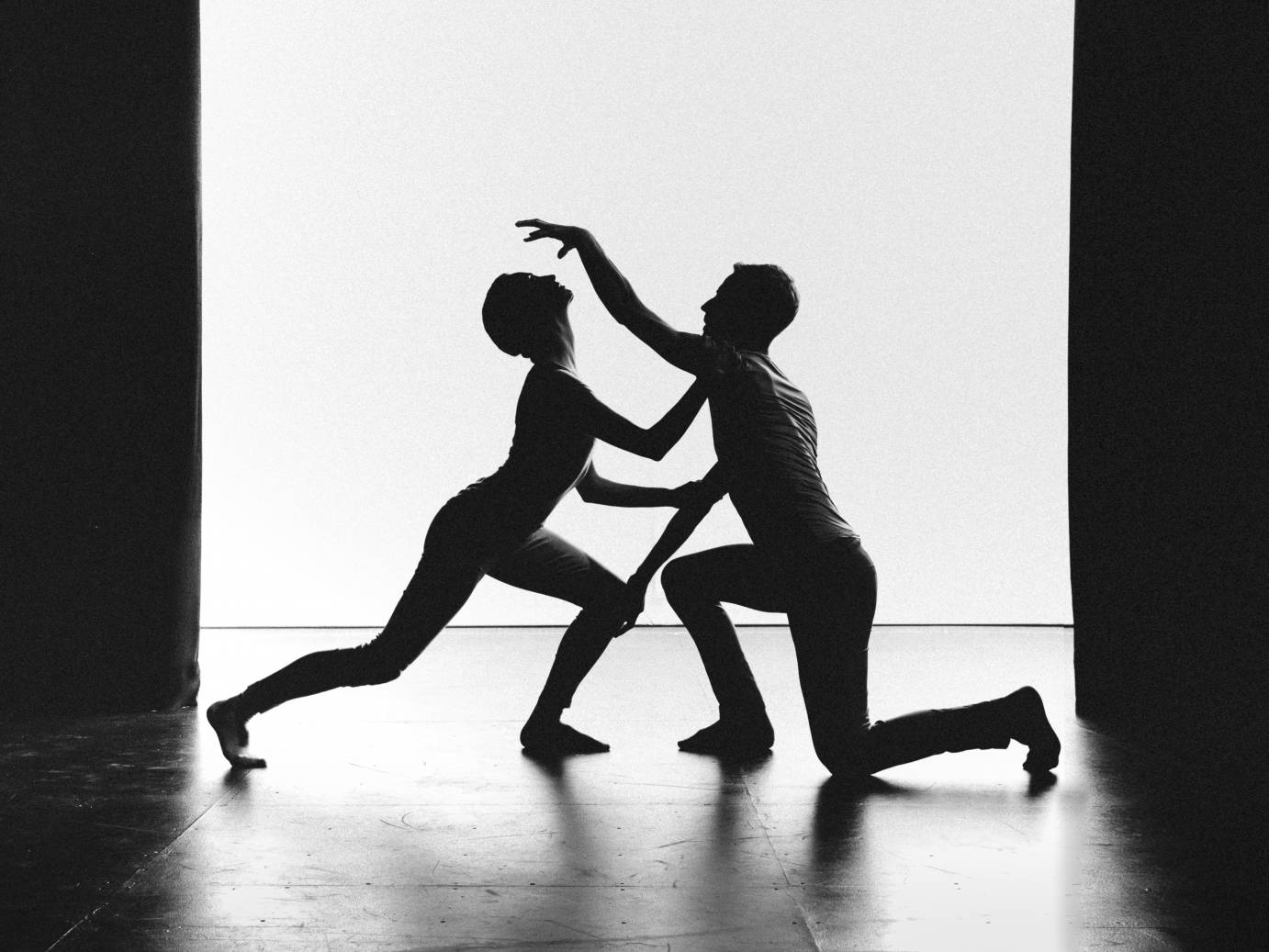 In profile, two dancers lunge facing each other as one lifts a hand above and the other lifts her lips
